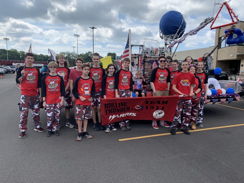 Team members dressed in red shirts and camo pants holding a Team 1511 banner in front of a parade float with robots on it