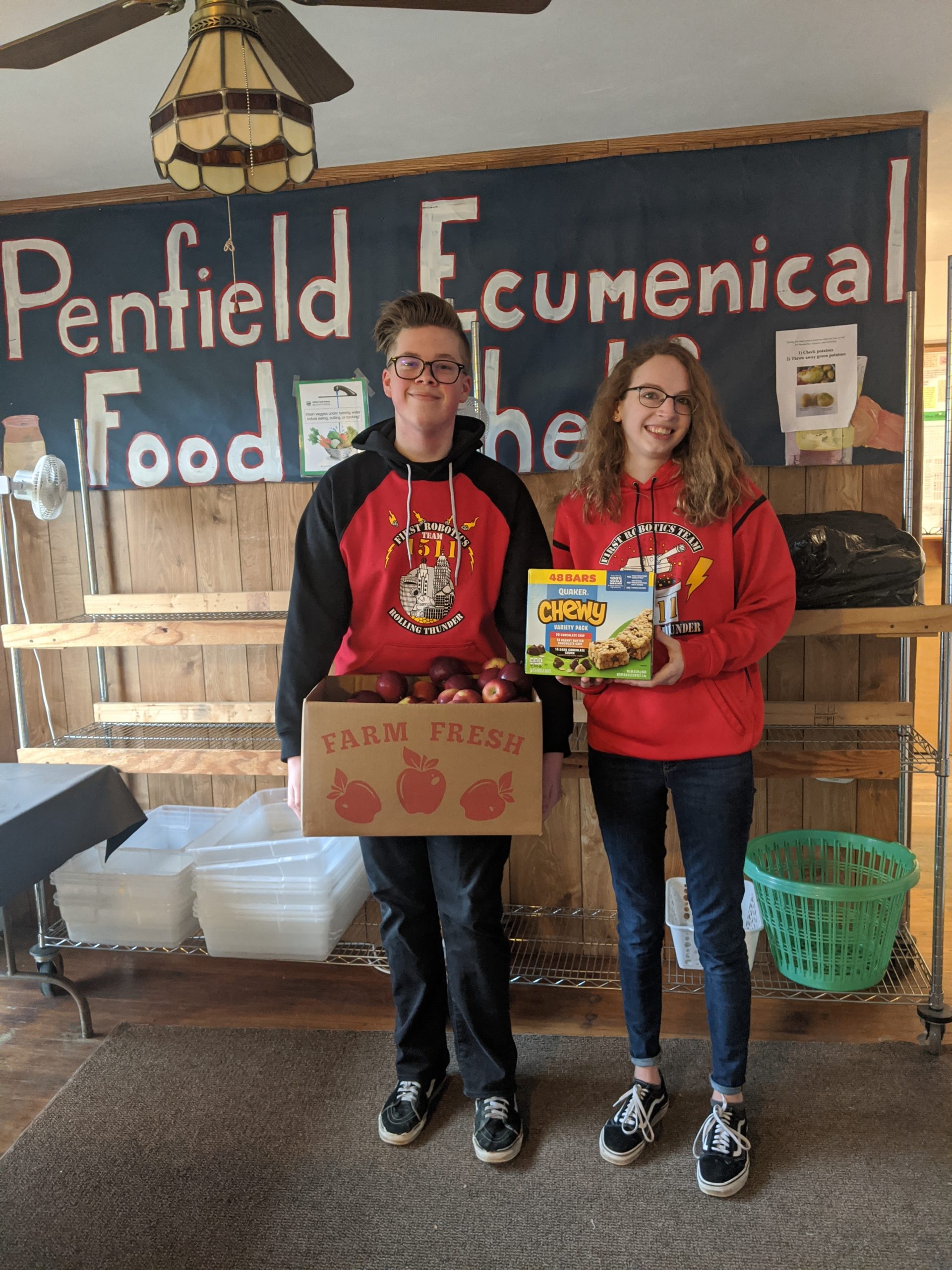 2 team members holding a box of apples and granola bars in front of Penfield Ecumenical Food Shelf sign