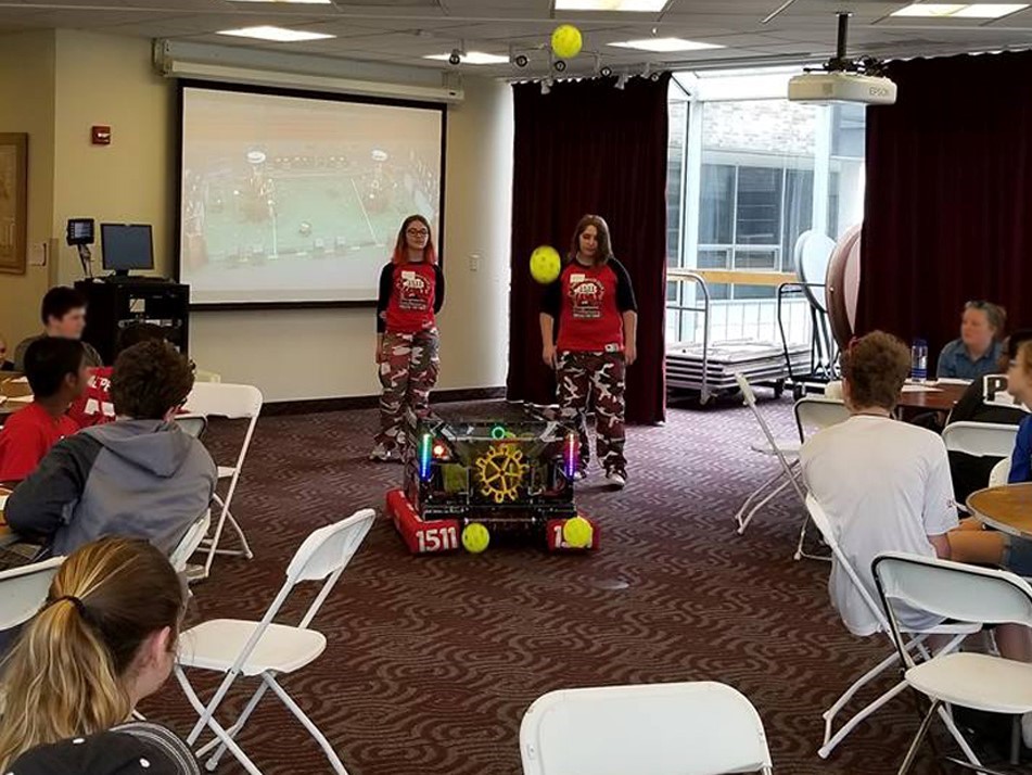 2 students with a robot shooting balls into the air while students site at tables watching at St. John Fisher College Science Exploration Day